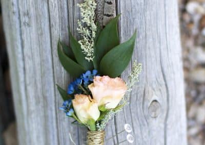 Grooms boutonniere that includes pink mini roses, blue wax flower, sage brush, pheasant feather, and greenery.