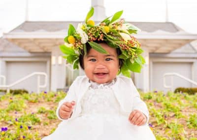 Baby girl with a flower crown with lots of greenery, white freesia, yellow billy balls, and pink pepper berries.