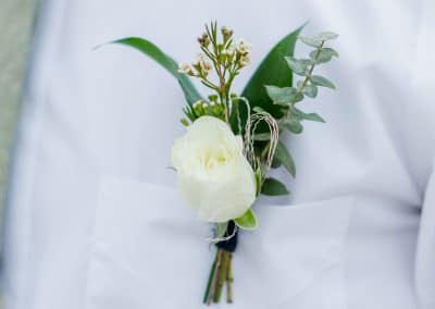 Boutonniere pinned on a white shirt that includes a ivory rose, wax flower, with a navy ribbon wrap.