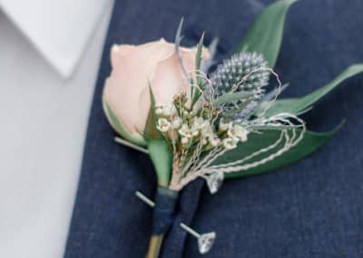 Boutonniere pinned on a jacket that includes a blush pink rose, thistle, wax flower, with a navy ribbon wrap.