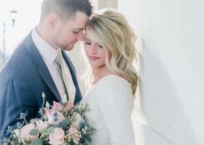 Bride and groom caressing while bride is holding and looking down at her bouquet that includes blush pink roses, thistle, and eucalyptus.